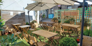 Sunny outdoor seating area with wooden tables, umbrellas and planted borders at Twisted Currant in Porthleven.