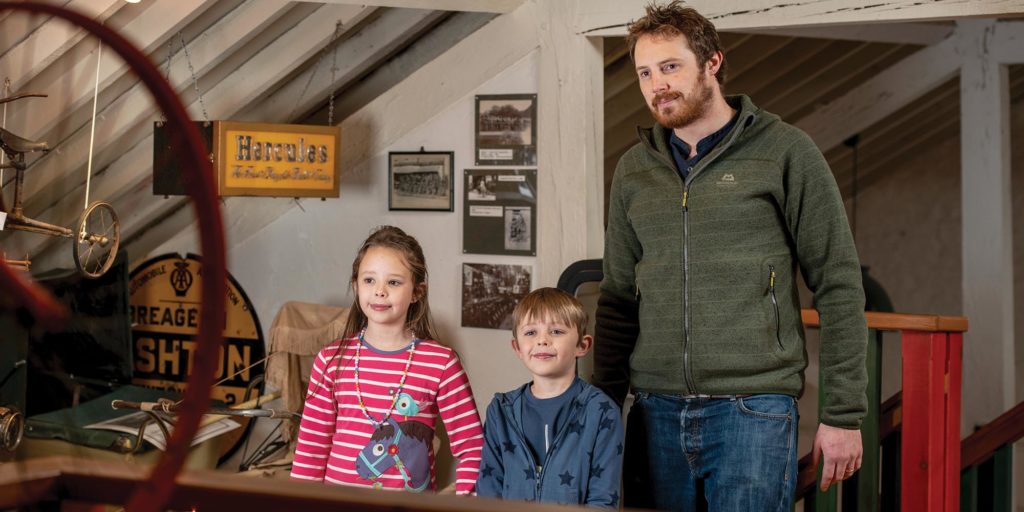 A man and two children looking at historic exhibits inside a museum, surrounded by vintage signs, photographs and machinery.