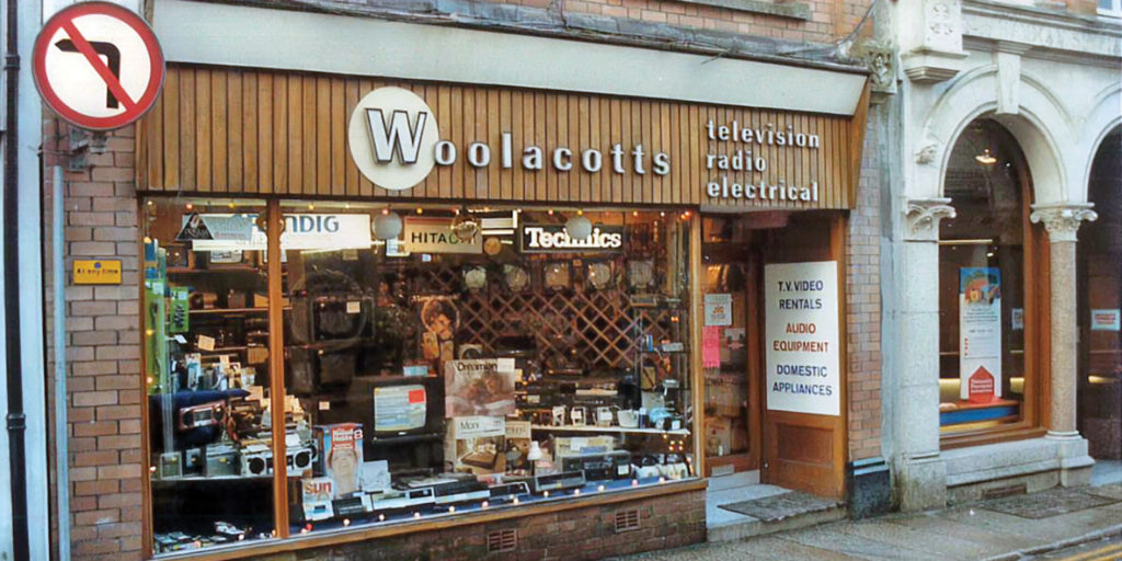 Historic exterior of the H.B.H. Woolacotts shop in Bude, with a red awning and electrical goods displayed in the windows.