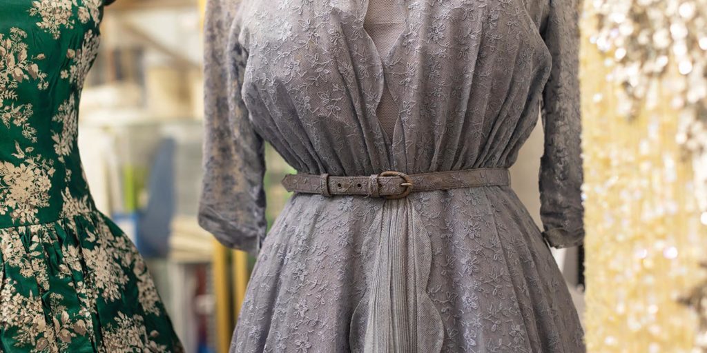 Close-up of vintage dresses on display, including a grey lace dress with a belt and a green floral dress beside it.