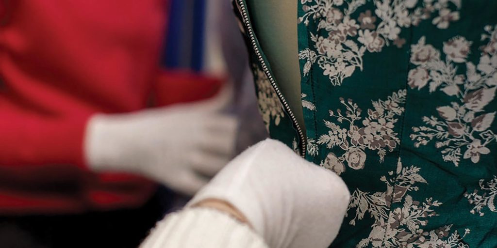 Close-up detail of a green floral dress on a mannequin, with blurred museum display items in the background.