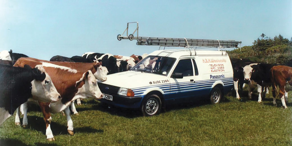 A vintage Woolacotts service van parked in a field, surrounded by cows.
