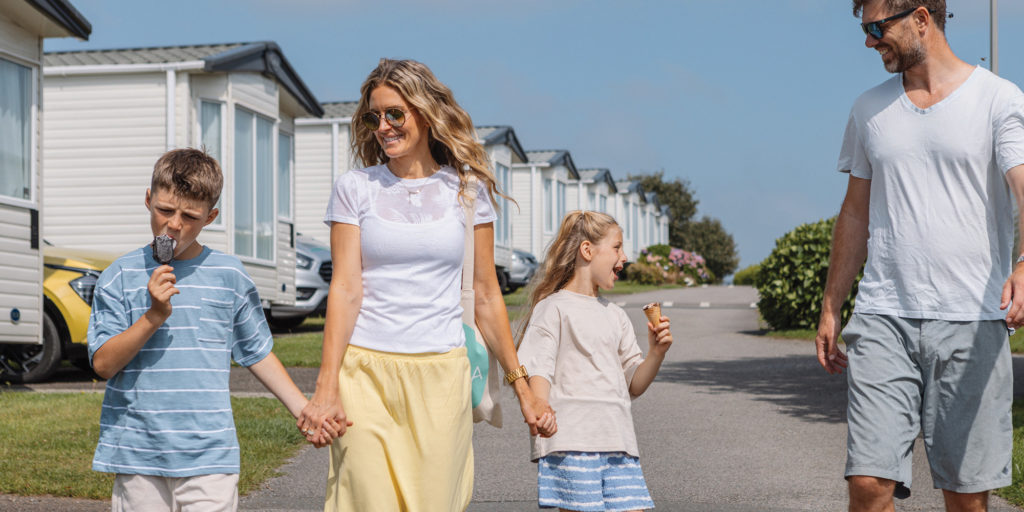 A family walking through a holiday park on a sunny day, with children eating ice creams beside rows of caravans.